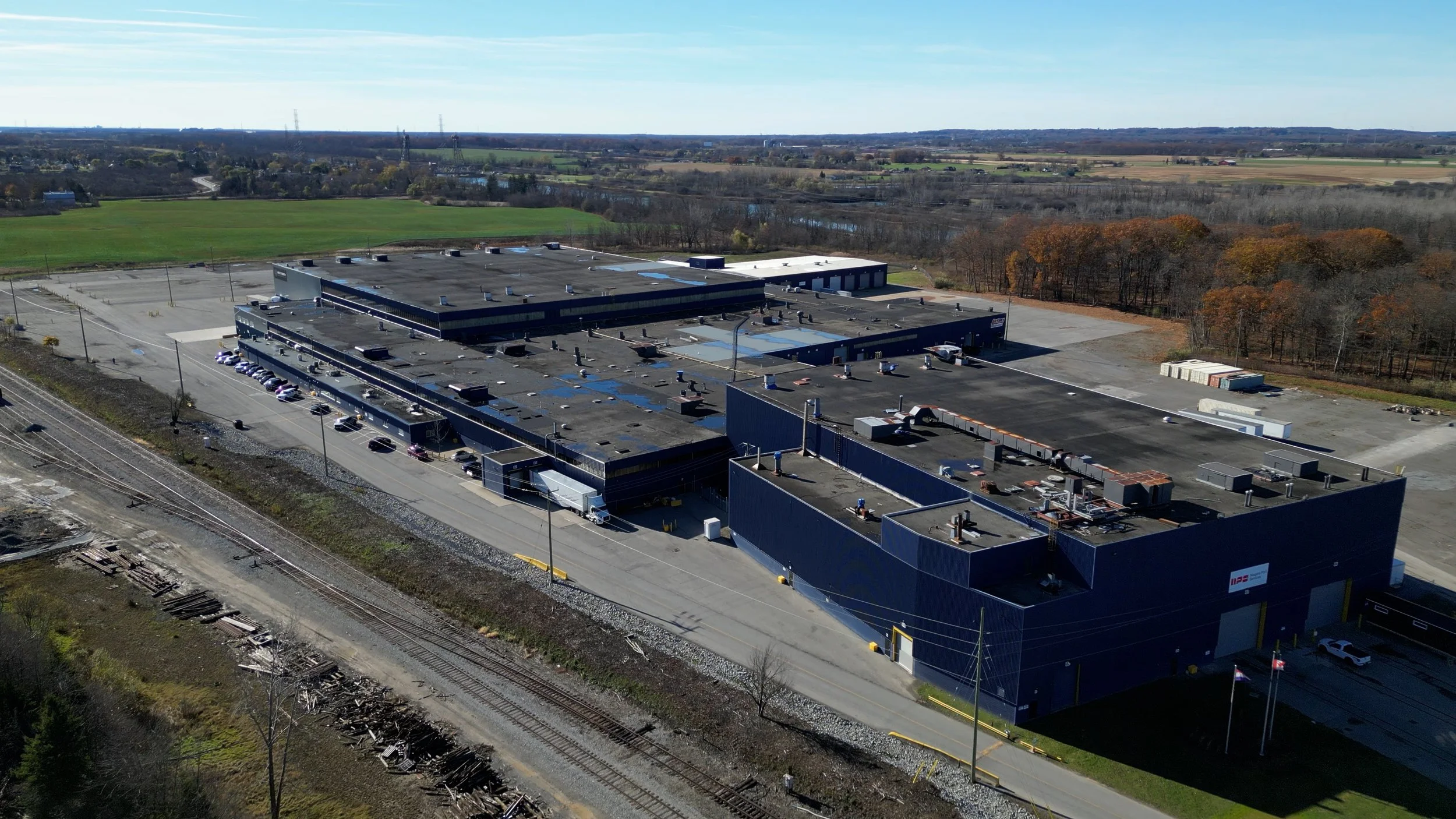 An aerial view of a large industrial warehouse surrounded by parking lots, with train tracks running nearby and wooded areas in the background.
