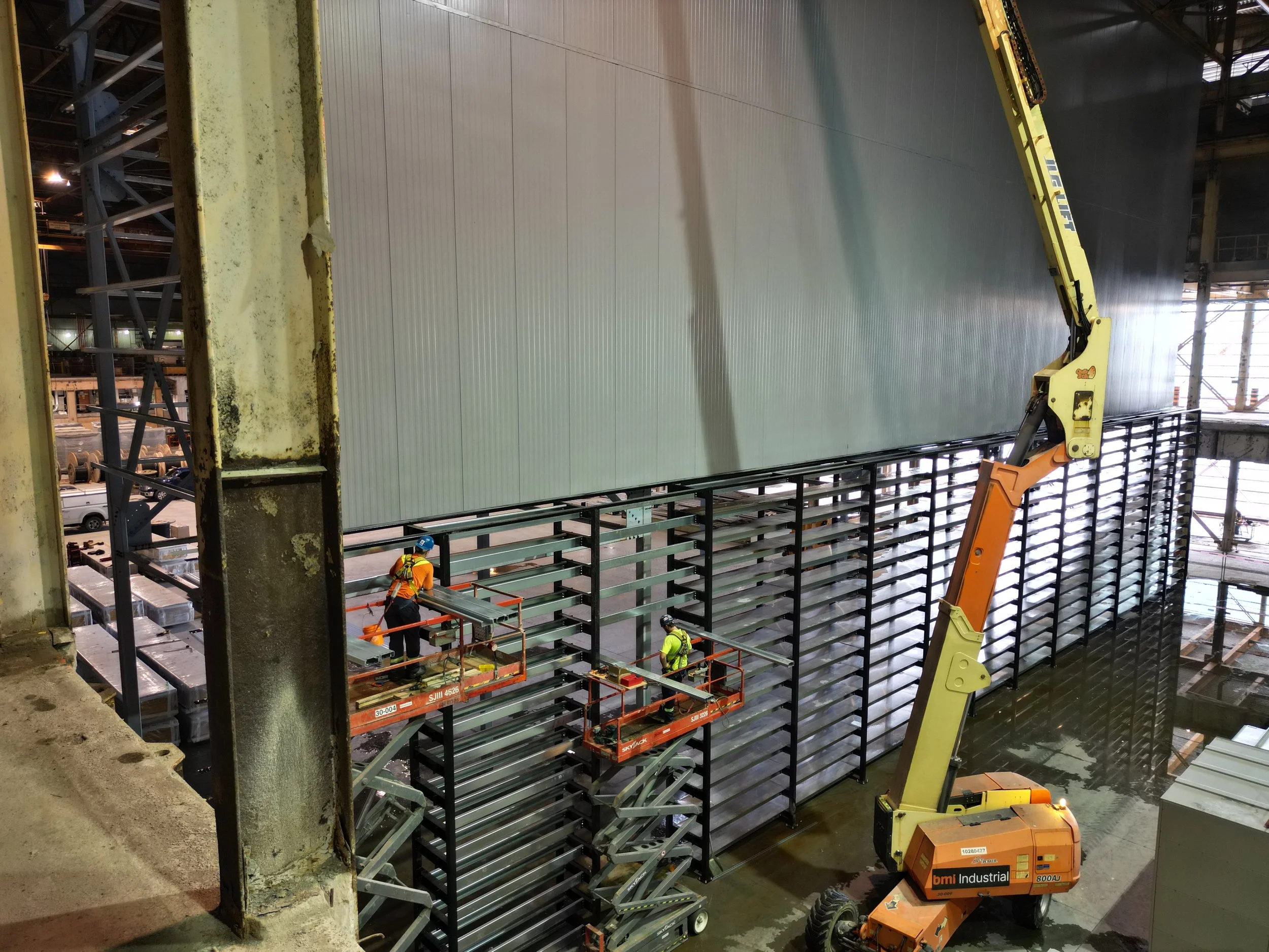 Construction workers in safety gear working on a large wall with a lift and a hydraulic arm inside a building under construction.