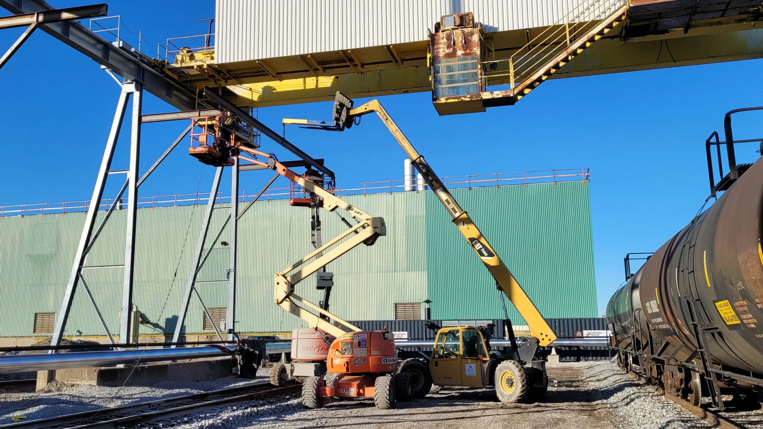 Workers on lift platforms inspecting and performing maintenance on a large industrial crane over railroad tracks, with train cars on the right and a green industrial building in the background.
