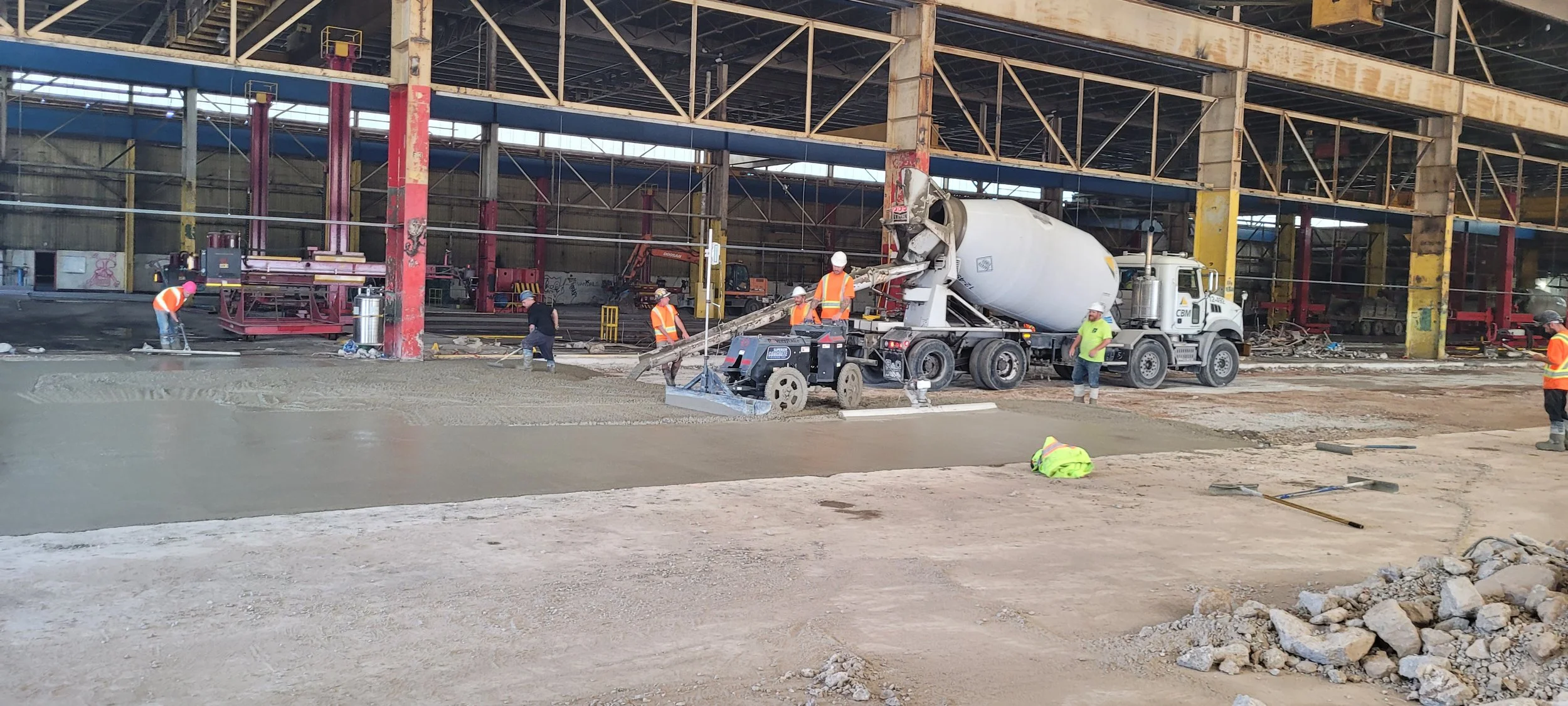 Construction workers pour concrete on a large indoor construction site, with a cement mixer truck and various construction equipment and materials.