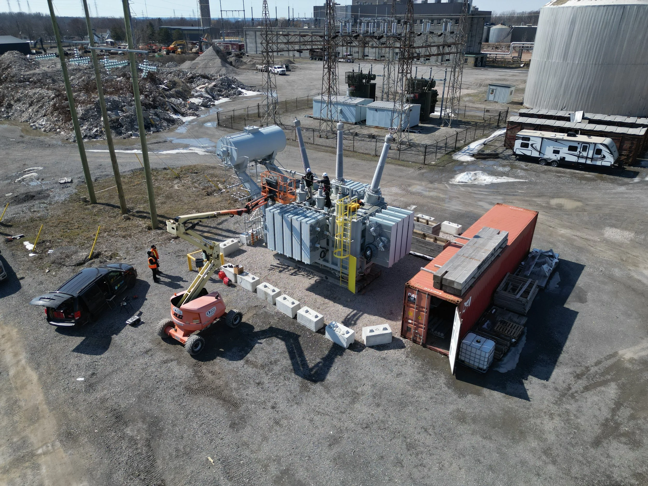 Construction site with a transformer, a crane lifting equipment, a red shipping container, a truck, and utility poles under construction.