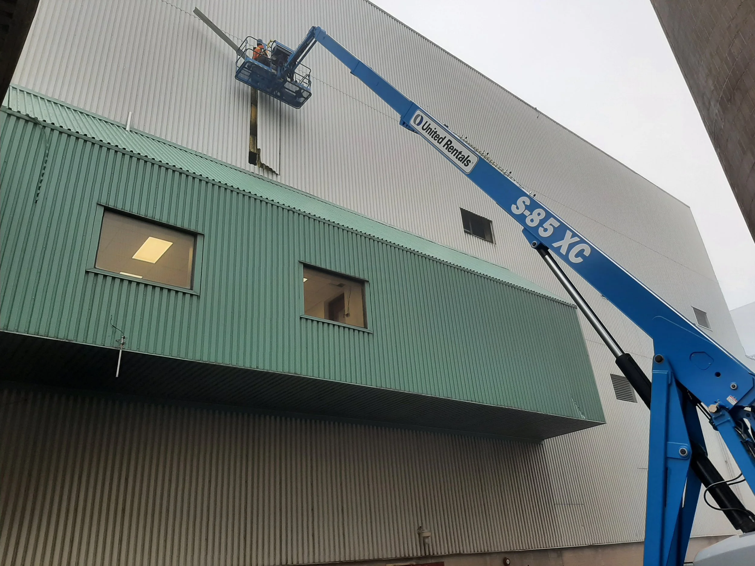 A blue crane lifts workers to repair a large hole in the side of a modern building with green and gray metal siding.