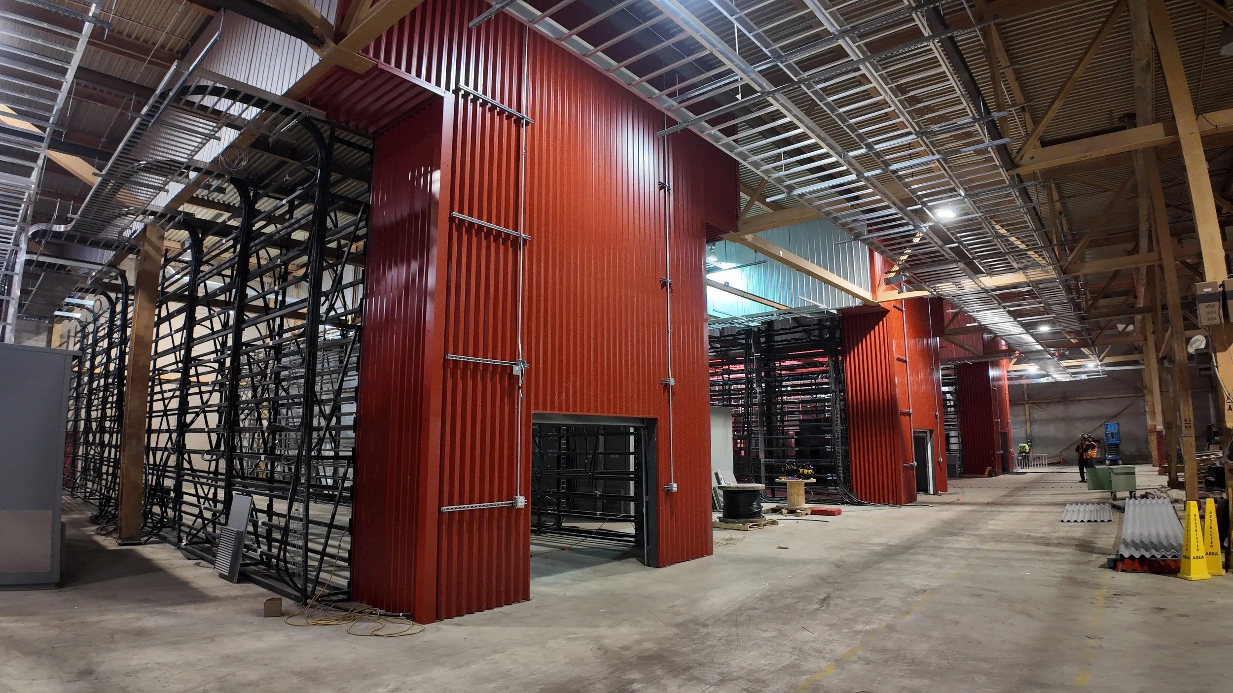 Construction site inside a large building with metal framing, red and black wall panels, and workers in the background.