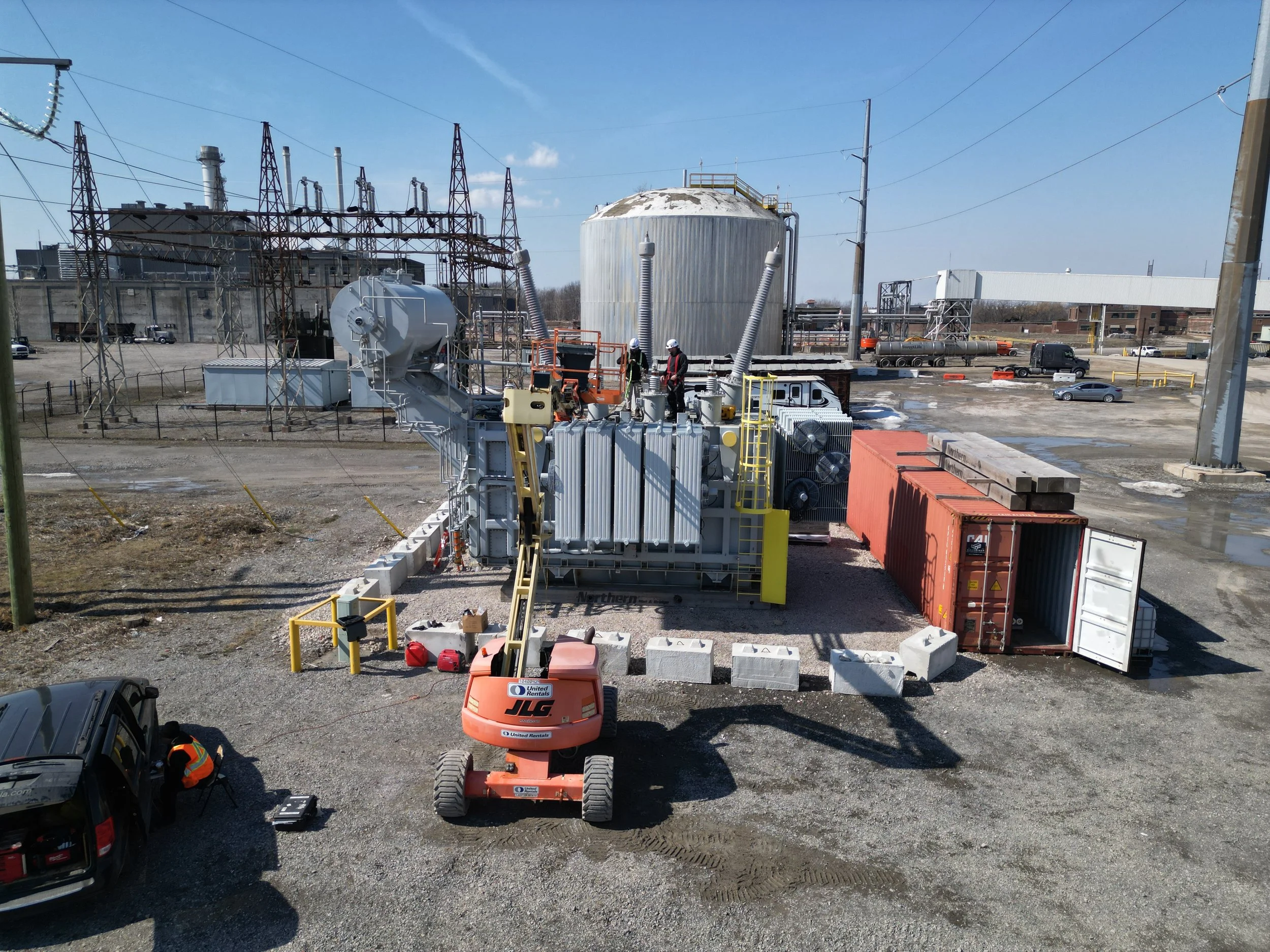 Construction workers operate heavy machinery at an industrial site with storage tanks, power lines, and a partially open shipping container.