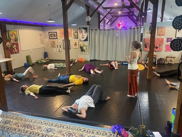 Group of people lying on the floor in a yoga or meditation class, with an instructor standing nearby, in a room decorated with artwork and soft purple lighting.