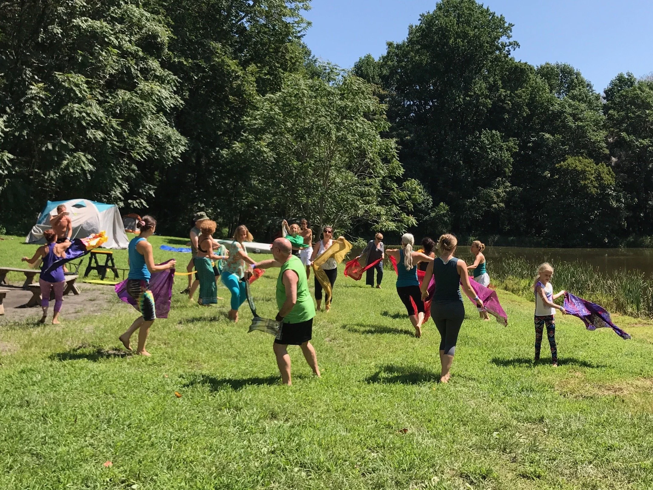 Group of people outdoors on a sunny day, participating in a collective activity by a lake, with tents and trees in the background.