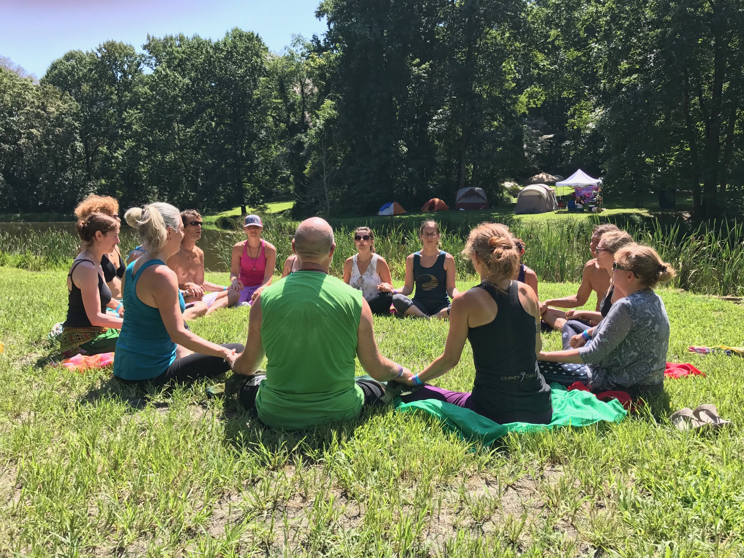 Group of people sitting in a circle on grass near a pond, holding hands, practicing yoga or meditation outdoors on a sunny day.