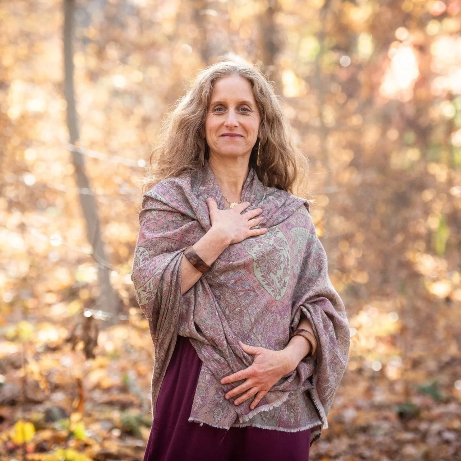 A woman with long, curly hair stands outdoors in an autumn forest, holding her hand over her chest and stomach area, wearing a patterned shawl and a maroon skirt, with fallen leaves in the background.