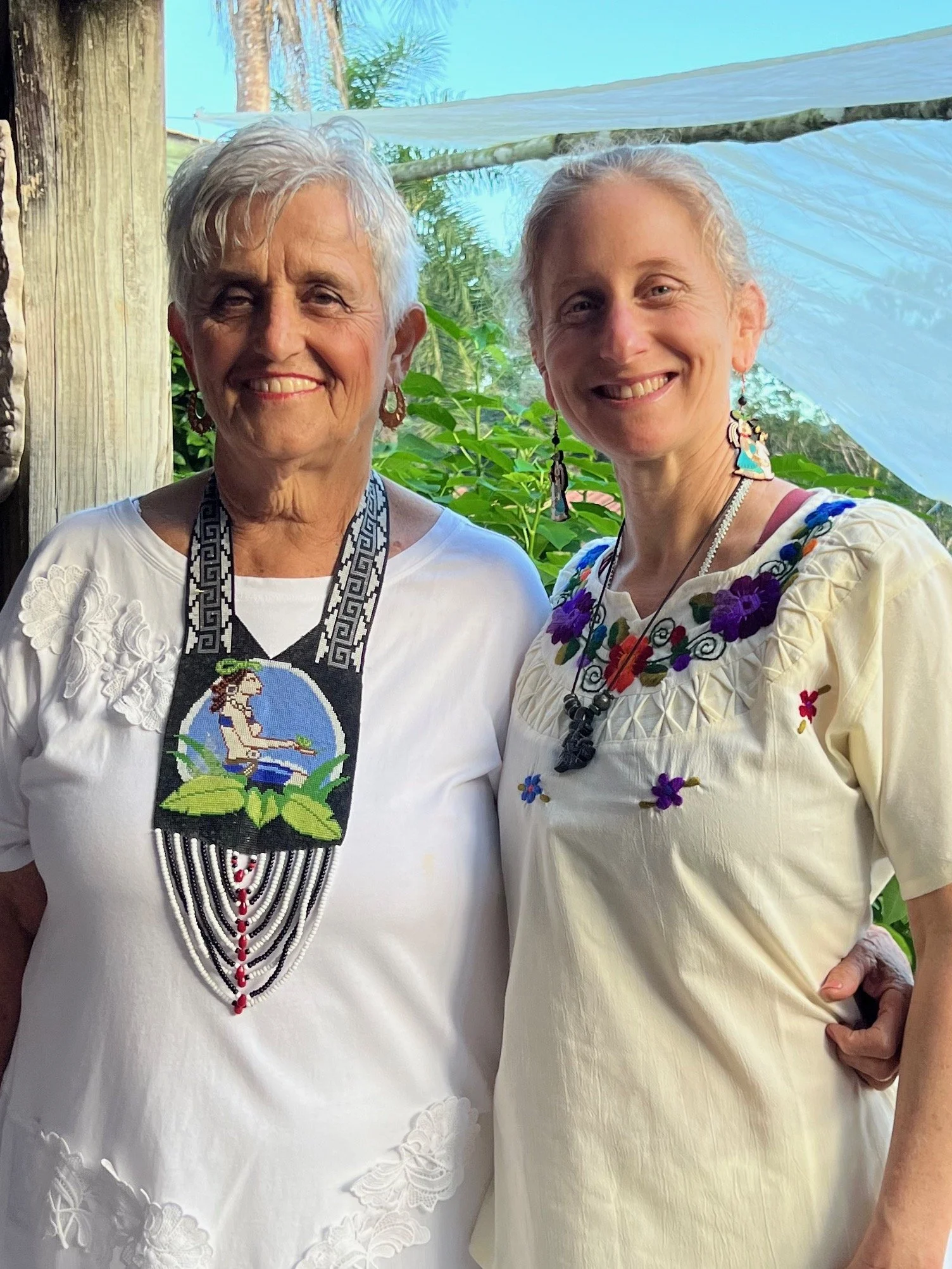 Two women smiling outdoors, wearing embroidered dresses and beaded necklaces, with green foliage and a blue sky in the background.