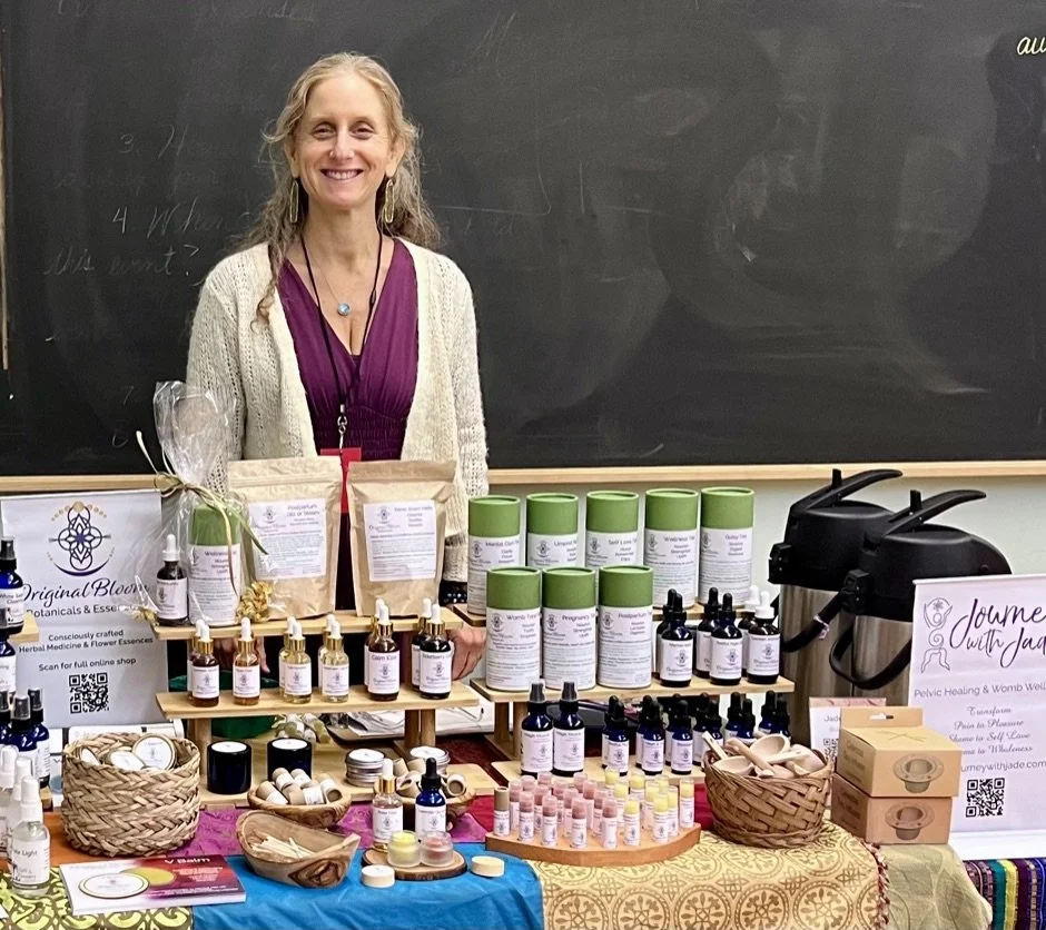 Woman standing behind a table with herbal products and essential oils, with a blackboard in the background.
