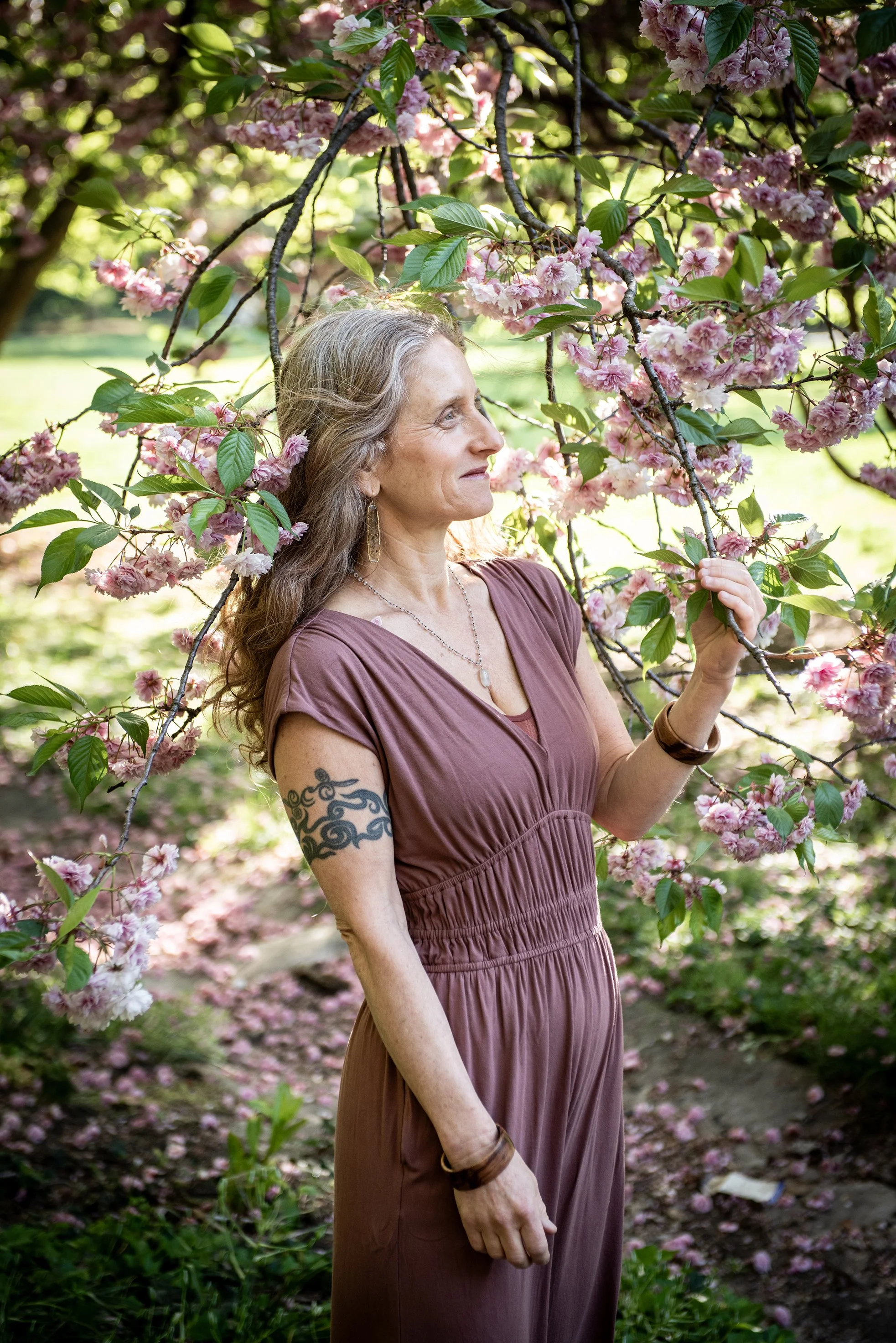A woman with long, curly gray hair standing outdoors among pink blossoming trees, holding a branch and smiling thoughtfully.