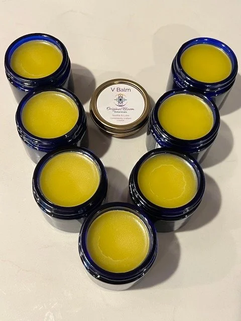 Seven blue jars filled with yellow balm arranged in a circle around a small tin labeled V Balm on a white surface.