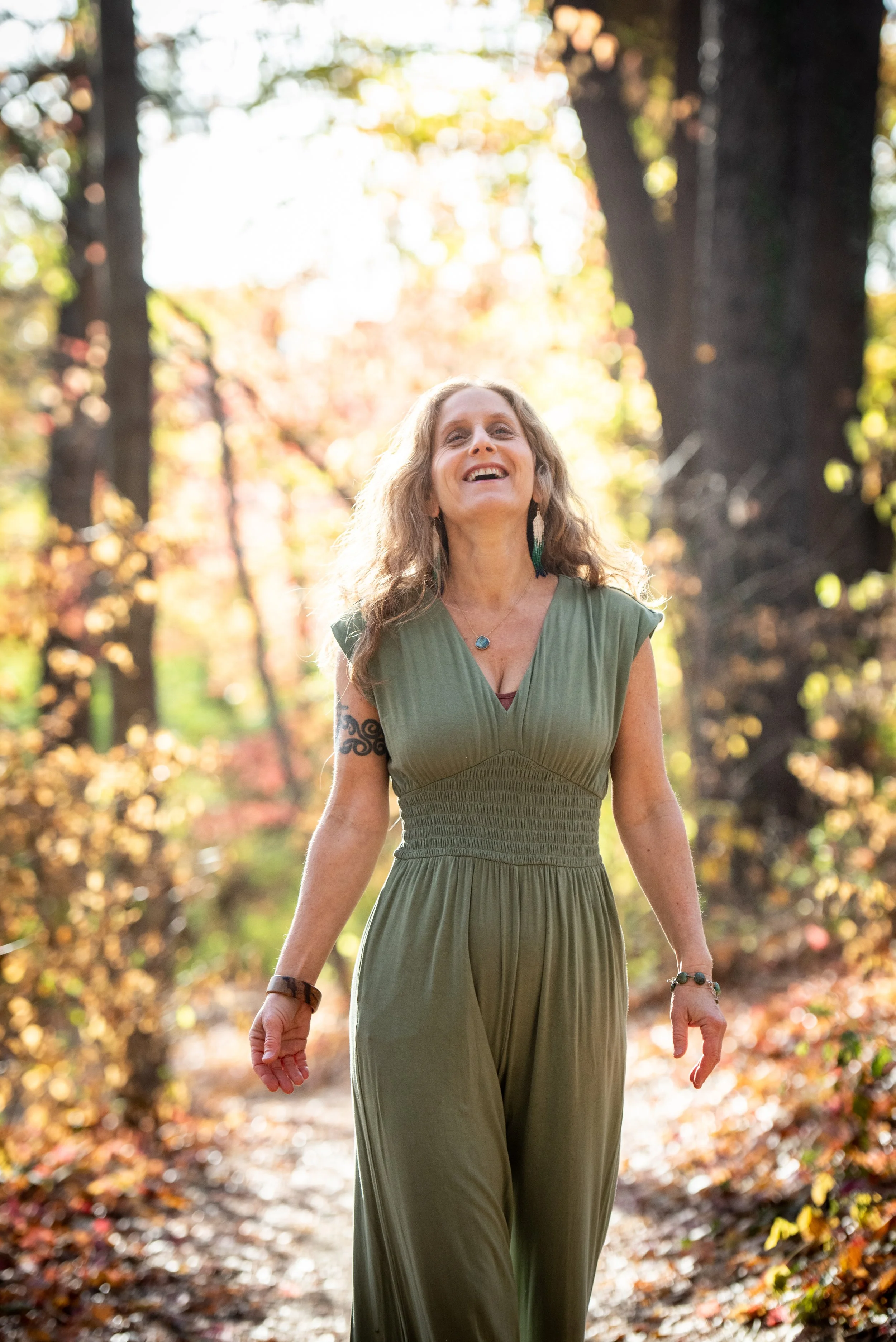 A woman with long curly hair smiling and walking outdoors in a forest during autumn, wearing a green dress.
