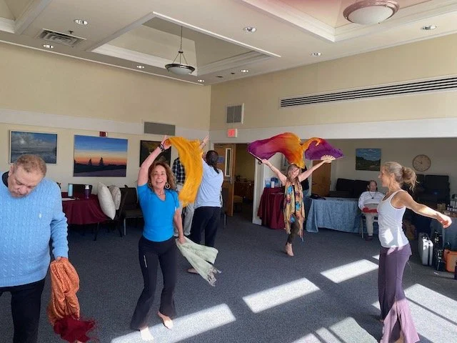 People dancing indoors, some holding colorful silk scarves, with paintings on the walls and tables in the background.