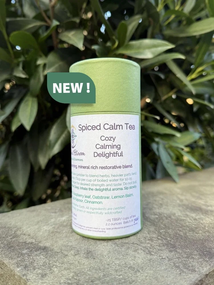 A green cylindrical canister of 'Spiced Calm Tea' with a label describing it as a cozy, calming, and delightful herbal tea blend, placed outdoors in front of green leafy bushes. The canister has a 'NEW!' label on it.