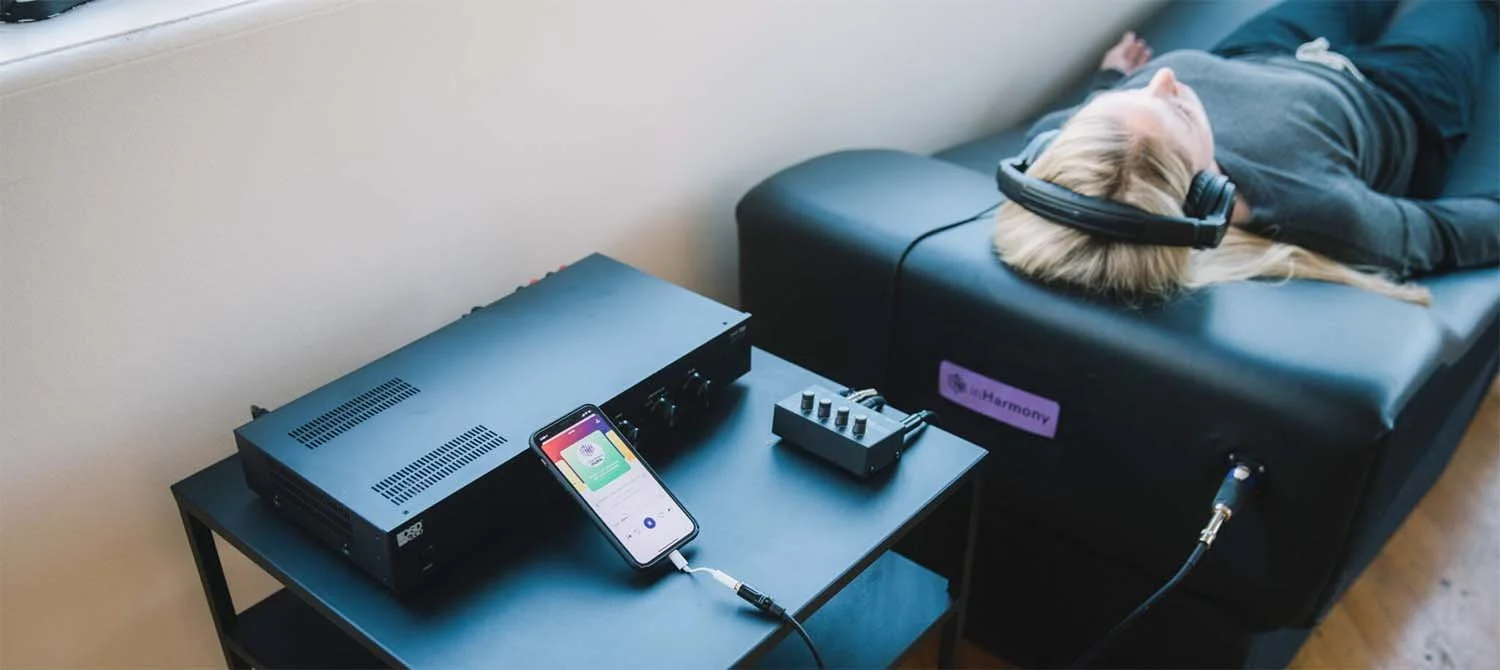 A person wearing headphones lies on a treatment table with a device attached to their head. Nearby, there is a black audio equipment on a small table connected to a smartphone, which displays an app interface. The setup appears to be for a neurological or auditory therapy session.
