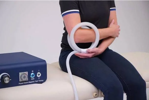 A person sitting on a medical examination table, holding a large ring connected to a pulsed electro-magnetic therapy device, used for muscle or pain treatment.