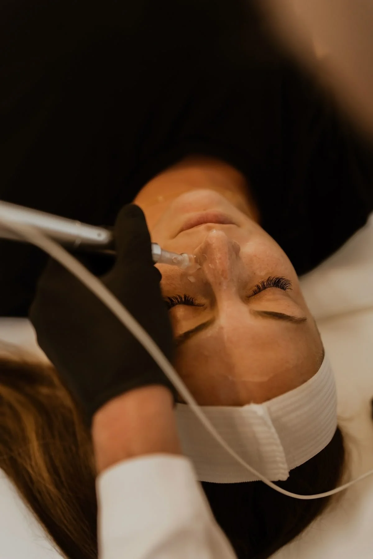 A person receives a facial injection while lying down with eyes closed, wearing a white headband and black top, with a technician administering the procedure with a syringe.