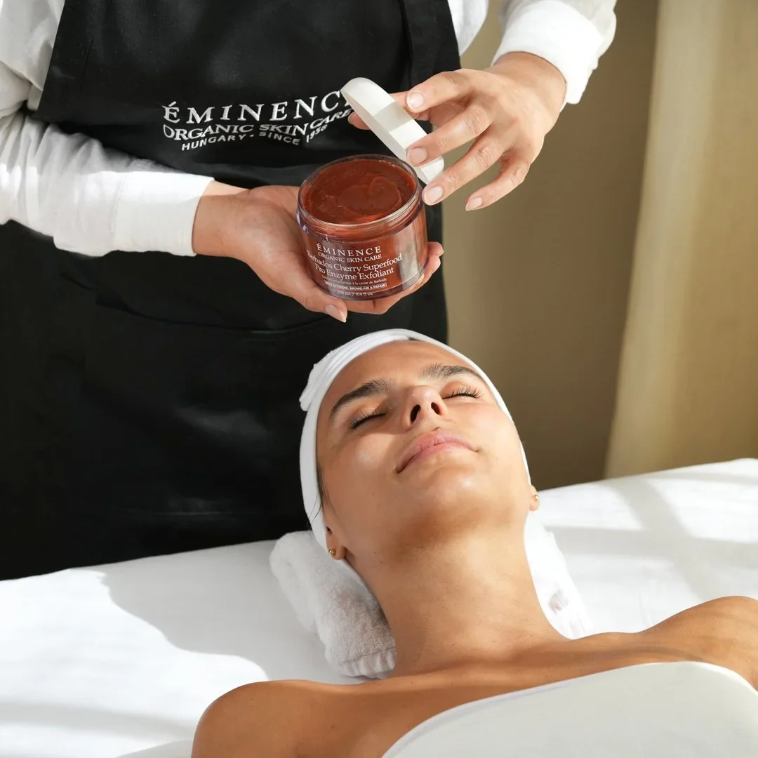 A woman receiving a skincare treatment while lying on a spa bed with her eyes closed, and someone applying a product from a jar labeled 'Éminence Organic Skin Care' to her face.