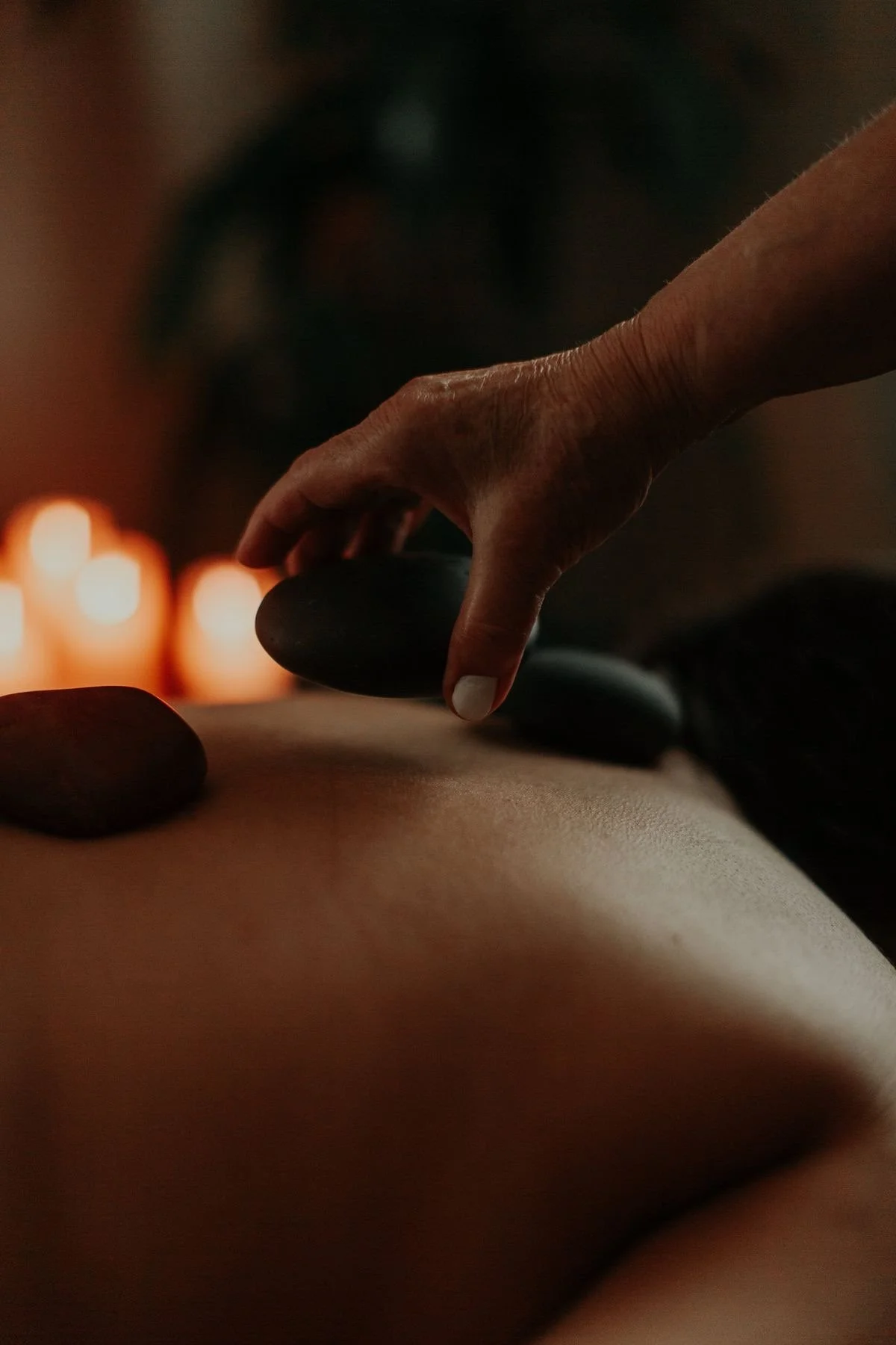 Close-up of a massage therapist's hand placing a black hot stone on a person's back during a massage in a dimly lit room with warm candlelight in the background.