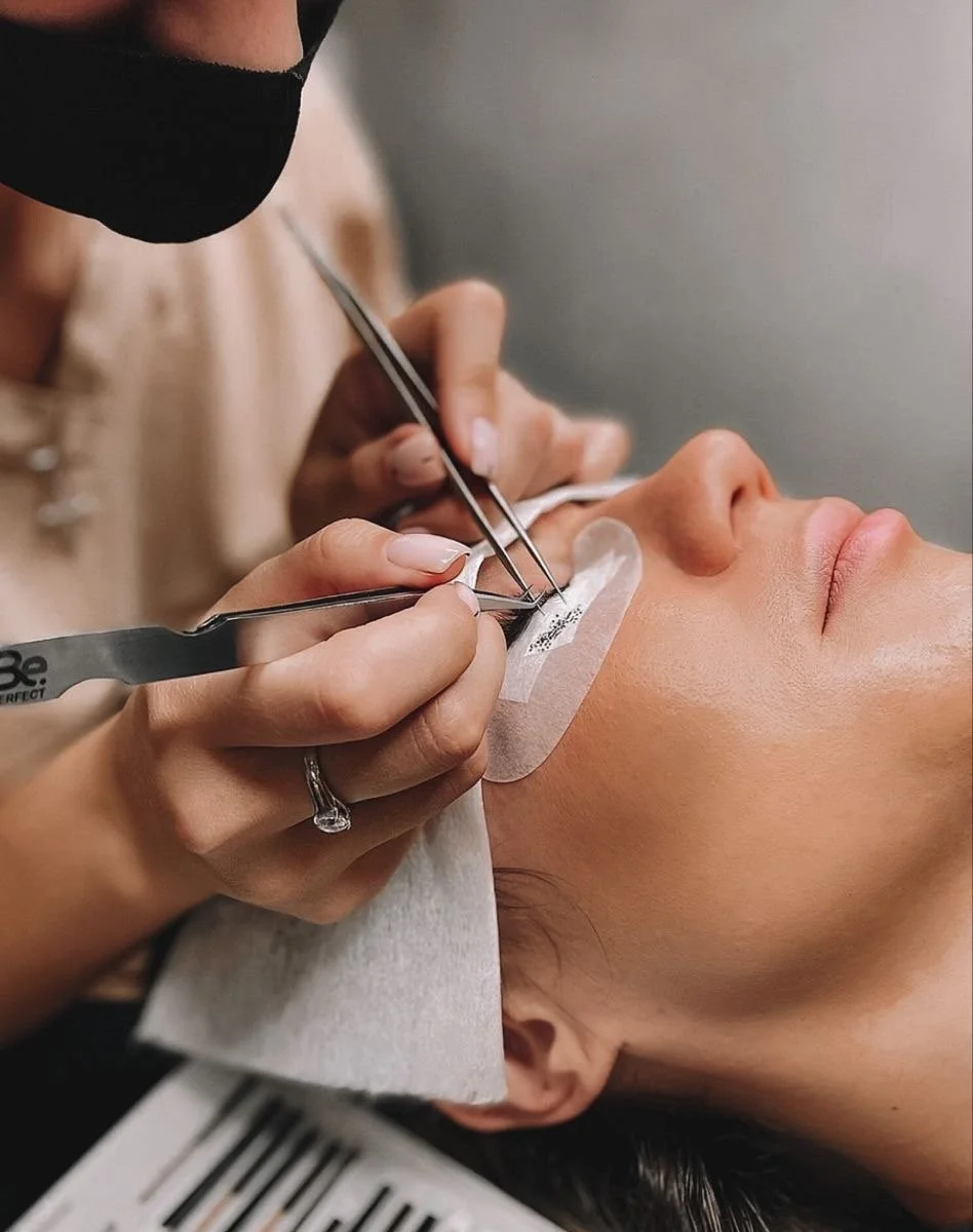 A person performing a cosmetic procedure on a woman's face, using dental tools to carefully work on her skin. The woman is lying down with her eyes closed, and there is a protective tape underneath her eye.