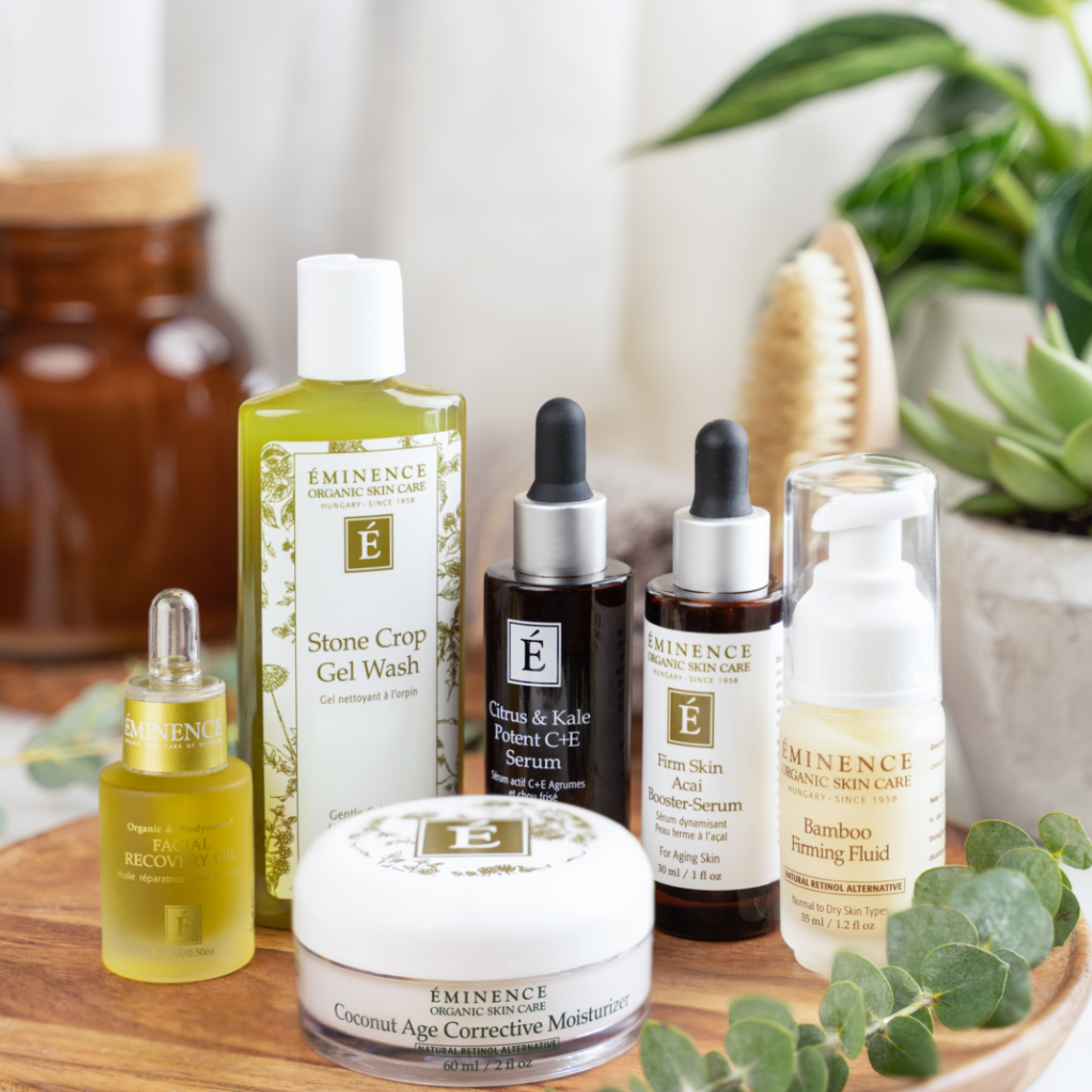 Collection of skincare products including gel wash, serum, booster, facial recovery oil, moisturizer, and bamboo firming fluid from Eminence Organic Skin Care, arranged on a wooden tray with plants in the background.