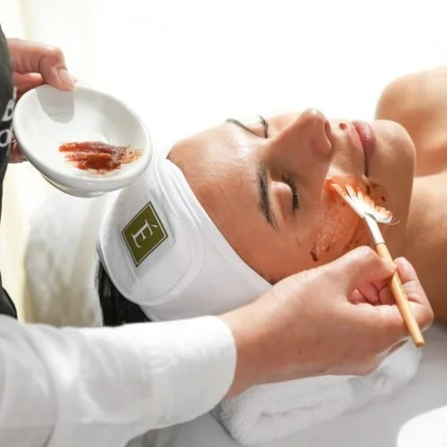 A woman receiving a facial treatment with a clay mask, lying with eyes closed, while a skincare professional applies the mask with a brush at the face spa in surprise.
