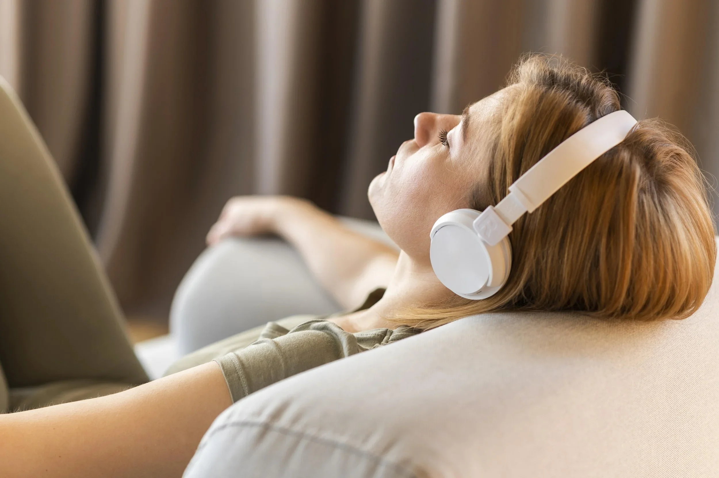 A woman with red hair is relaxing on a beige couch at a spa in Surprise, AZ, wearing white headphones, with her eyes closed and head tilted back.