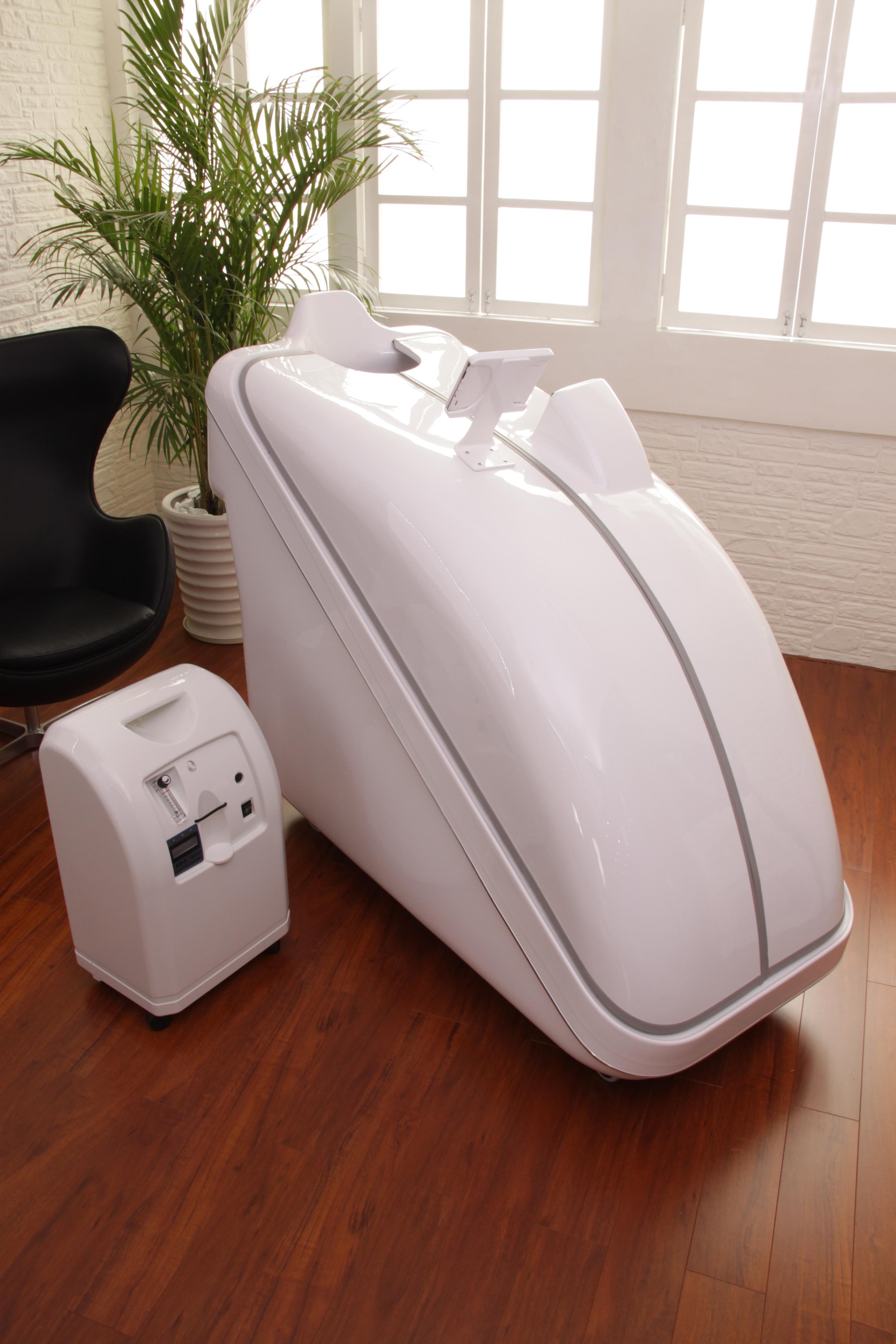 A white medical or wellness pod used for full body cleansing, with a screen on top, situated in a room with wooden flooring, a black chair, a large green plant, and a multi-pane window.