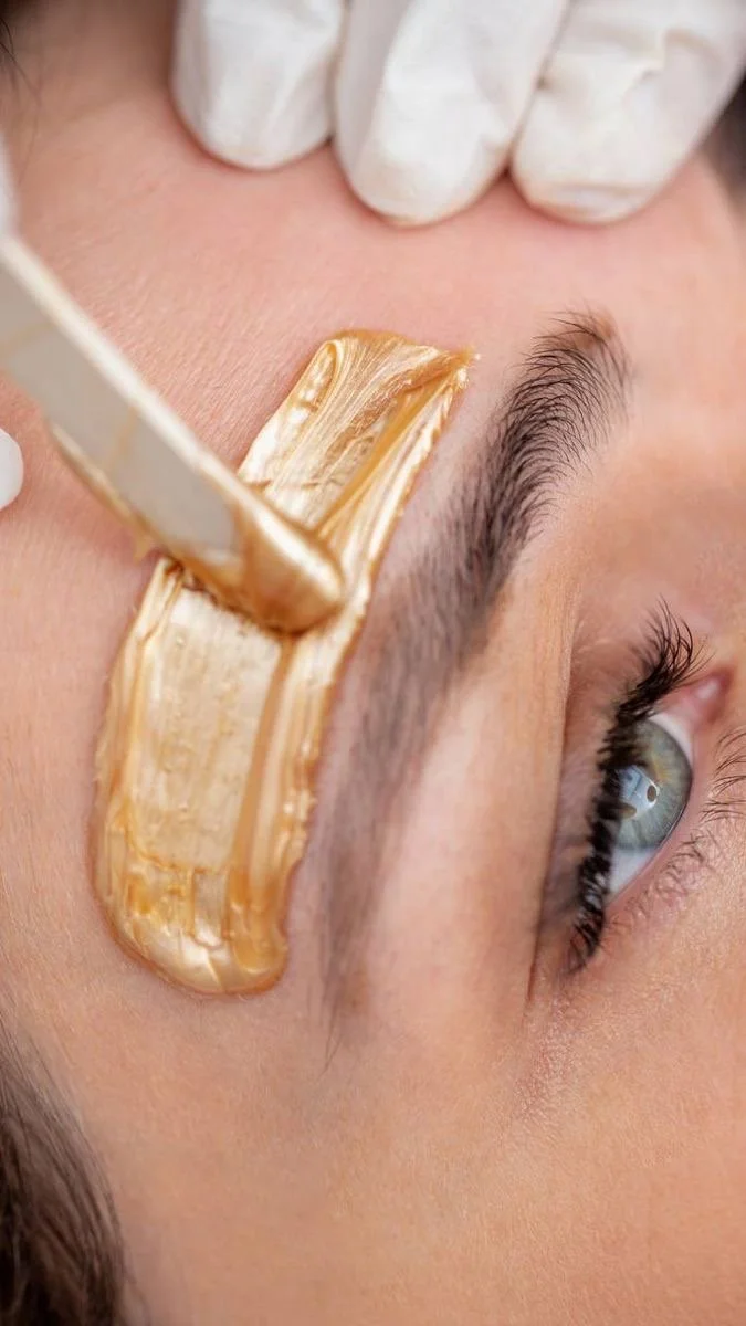 Close-up of a person's forehead and eye as a beauty professional applies a peel-off facial mask with a spatula.
