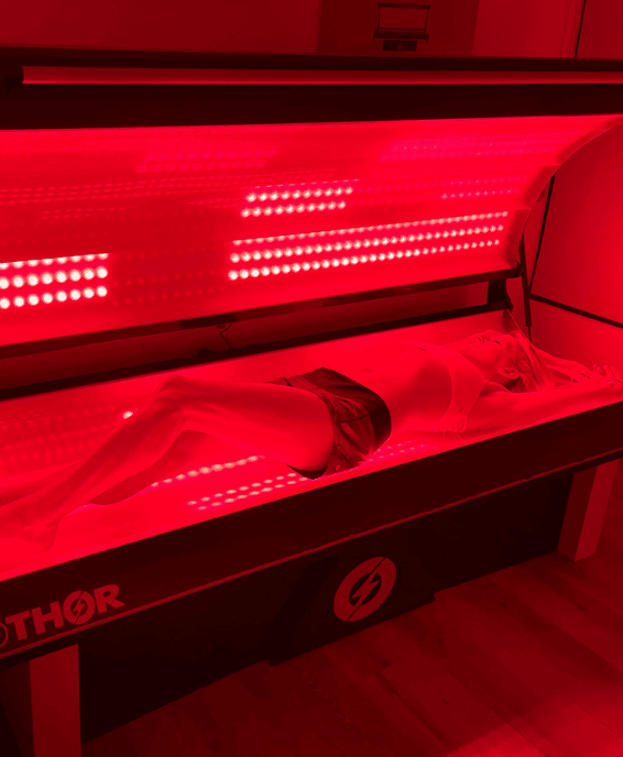Person lying inside a red light therapy bed with a glowing red light array above.