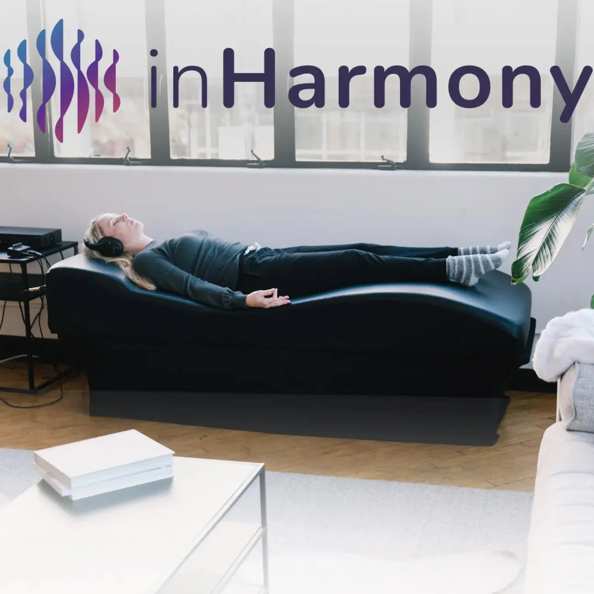 A woman with headphones relaxing on a black massage bed in a modern room with large windows, a plant, white furniture, and a logo that says 'inHarmony' on the window.