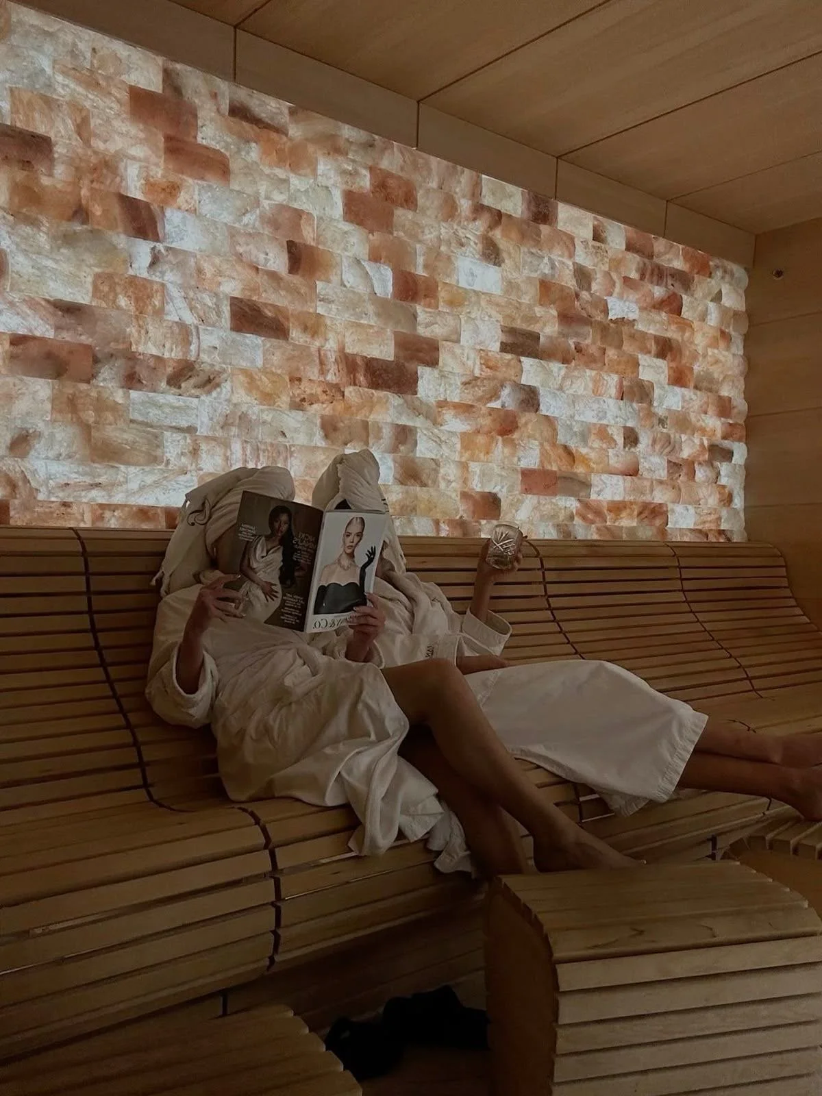 Person lying on a wooden slat lounge chair, wrapped in a white robe, holding a magazine with a female model on the cover in one hand and a glass in the other hand. The person is reading the magazine in a room with a brick wall and wooden ceiling.