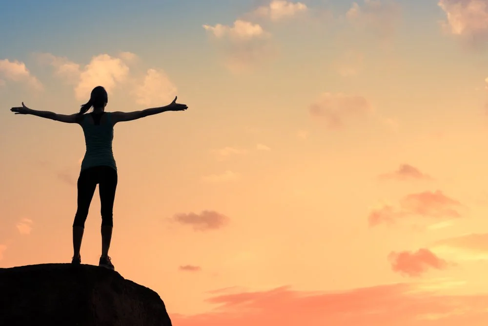 Silhouette of a woman standing on a rock with arms outstretched against a colorful sunset sky, representing a natural health coach.