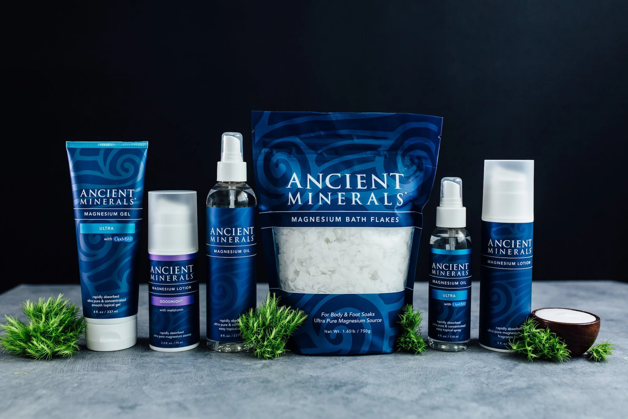 A collection of Ancient Minerals skincare products including magnesium gel, lotion, oil, bath flakes, and moisturizer, arranged on a gray surface with green moss and a halved coconut.