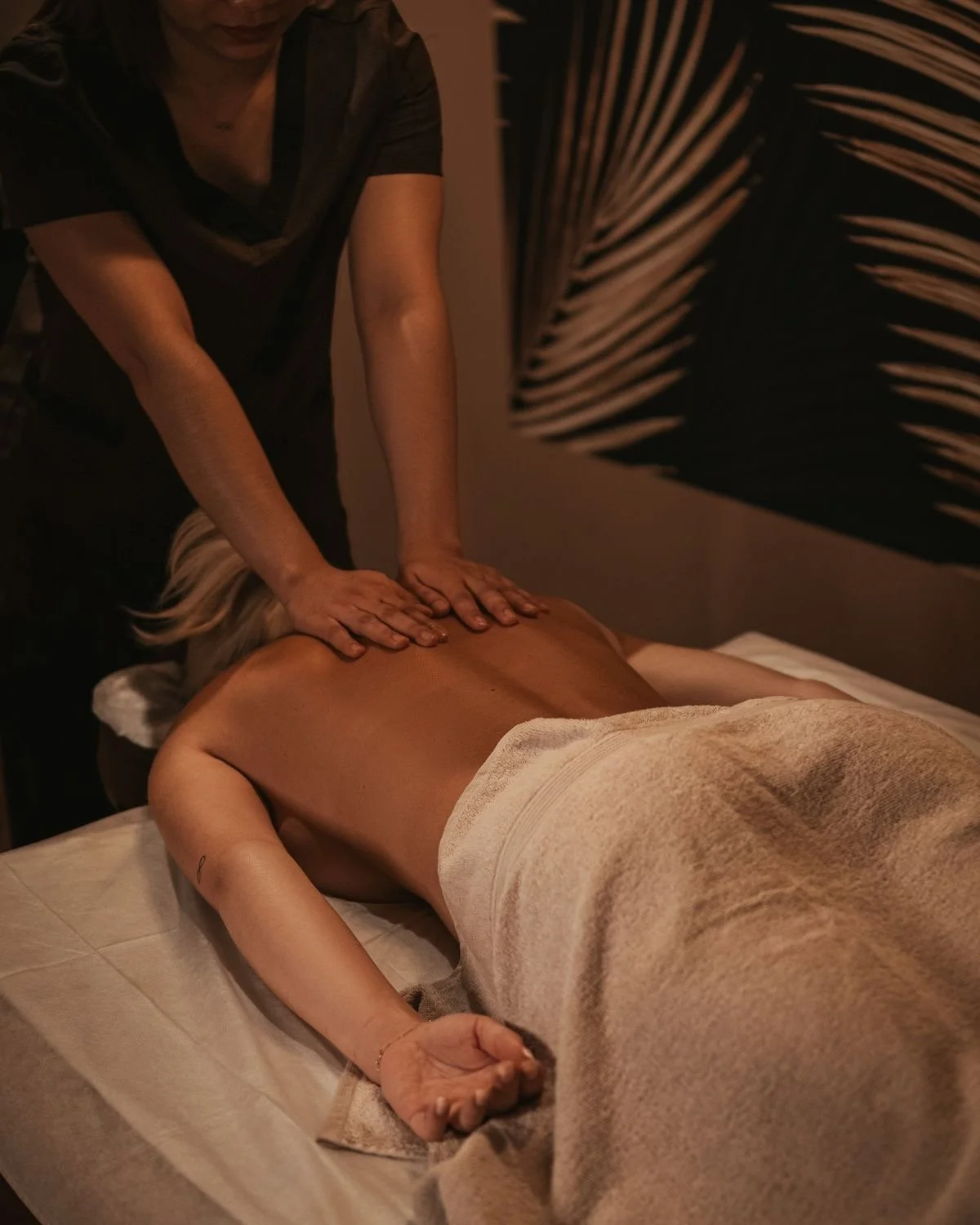 Person receiving a massage in a dimly lit room, lying face down on a massage table with a towel covering lower body, while the therapist applies pressure to their back.