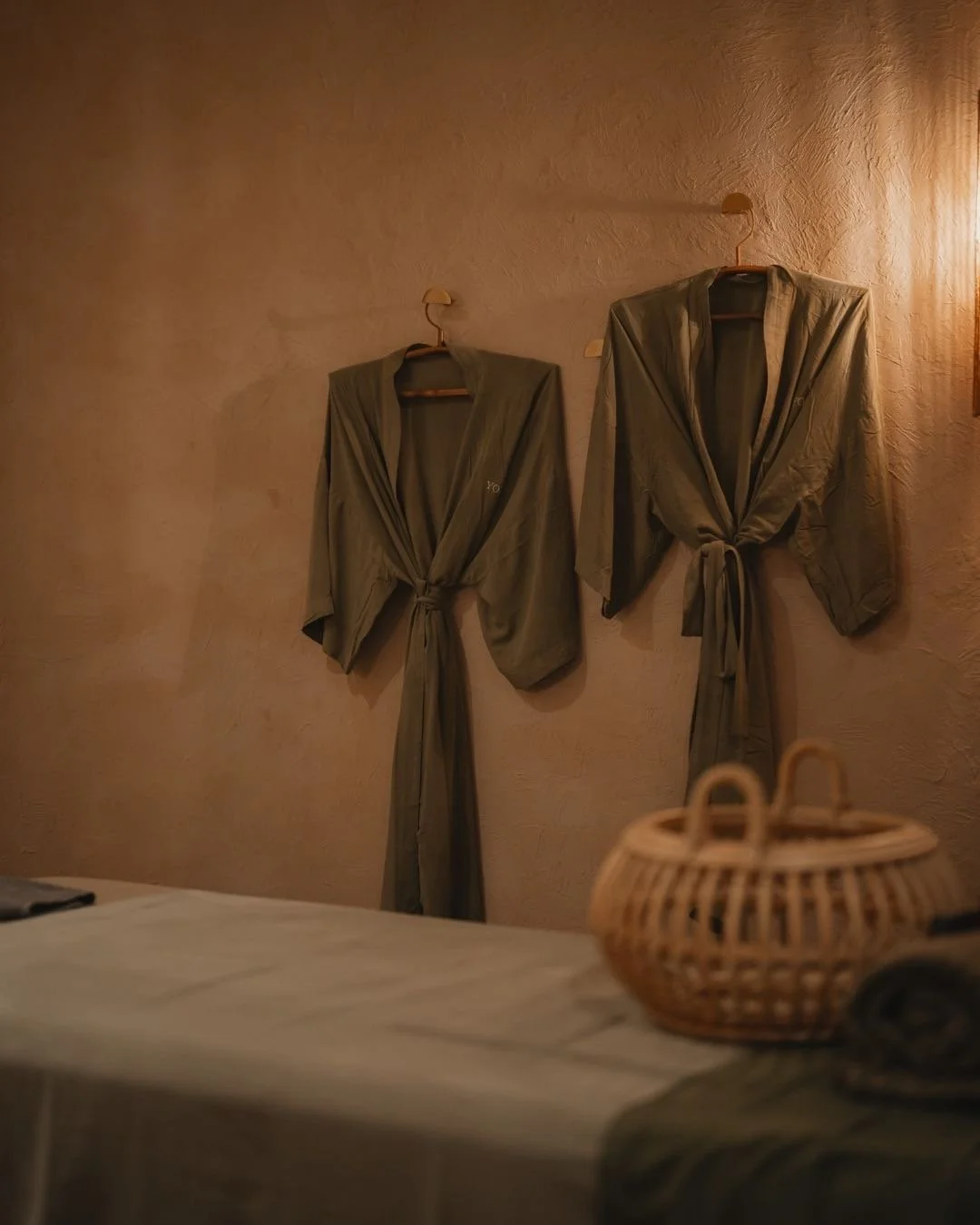 Two beige robes hanging on hooks on a textured wall in a dimly lit room.