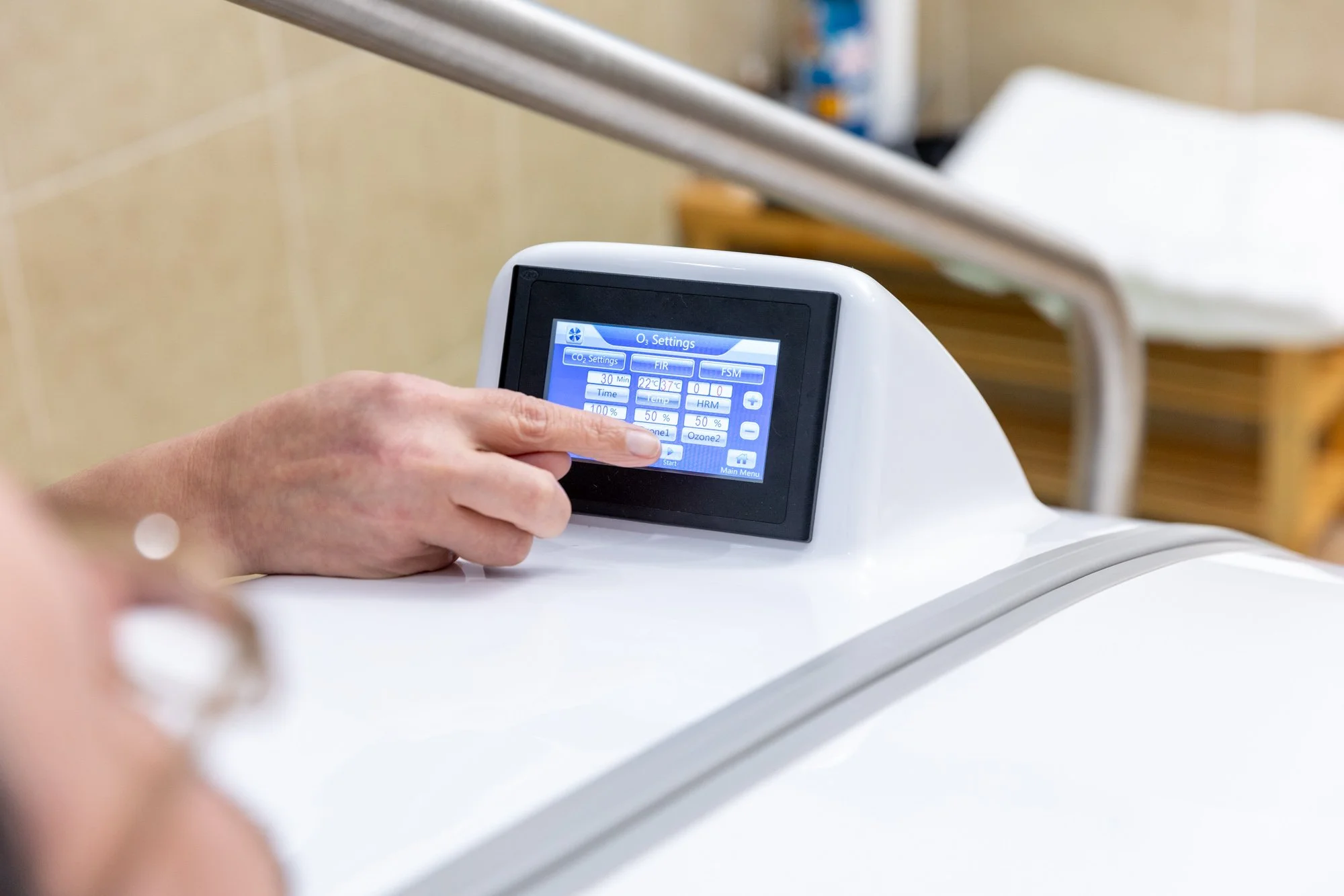 Close-up of a person's hand touching a touchscreen control panel on a medical device, with hospital equipment in the background.