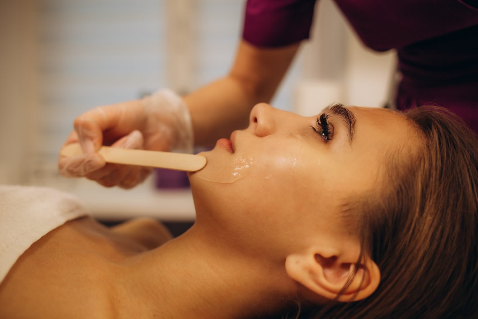 A woman receiving a facial treatment with a clay mask, lying with eyes closed, while a skincare professional applies the mask with a brush at the face spa in surprise.