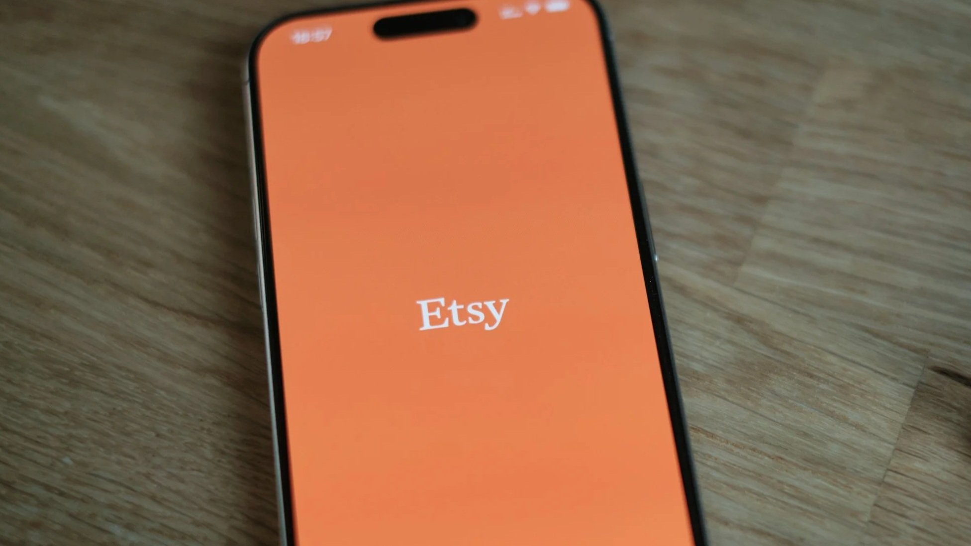 Smartphone displaying the Etsy app logo on an orange screen placed on a wooden surface.
