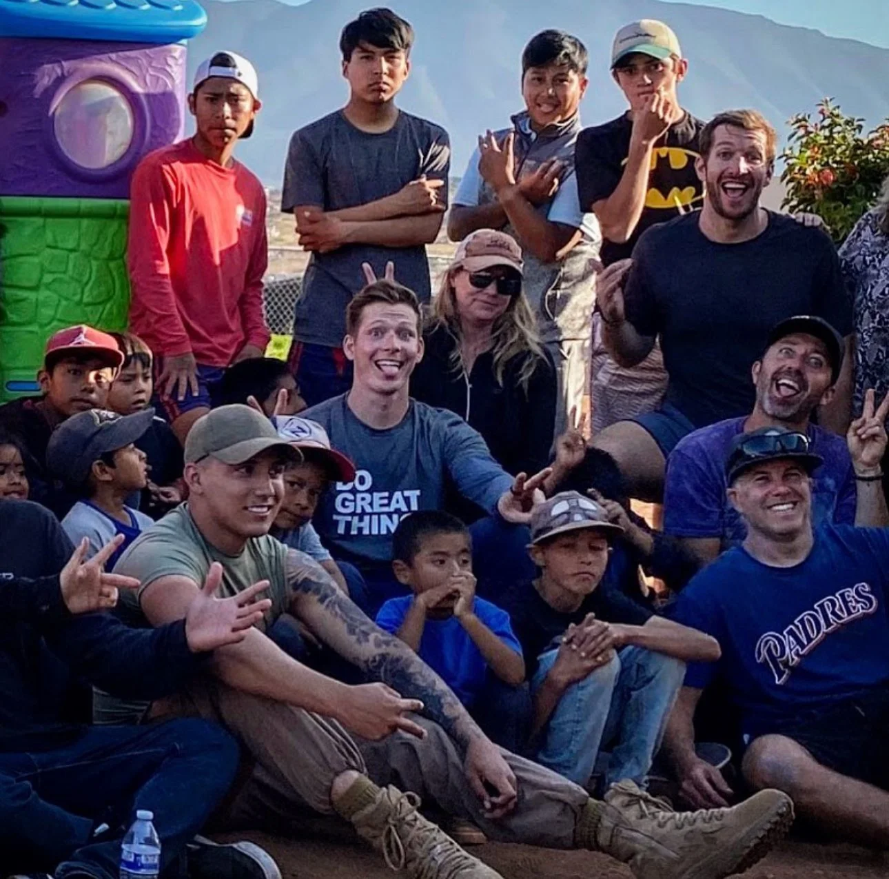 A group of children and adults at an outdoor event, some making peace signs and funny faces, with mountains in the background and colorful decorations around.