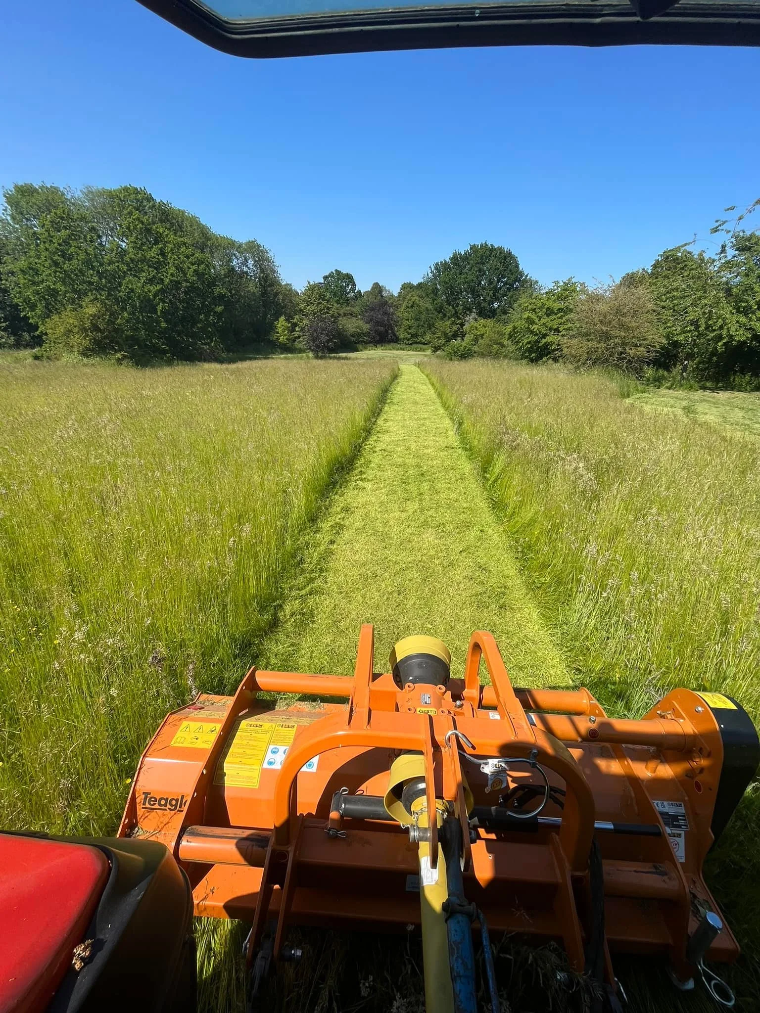 A view from a tractor in a field with grass and trees, showing a straight grassy path ahead under a clear blue sky.