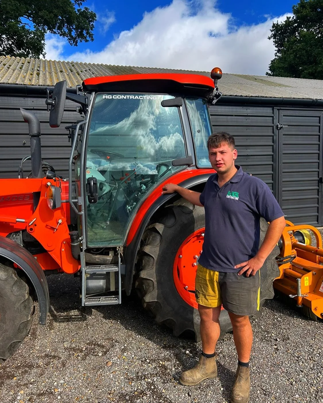 A young man in a navy blue polo shirt and shorts standing beside an orange tractor with large black tires; the man rests his hand on the tractor's cabin. The background includes a gray barn wall and a partly cloudy sky.
