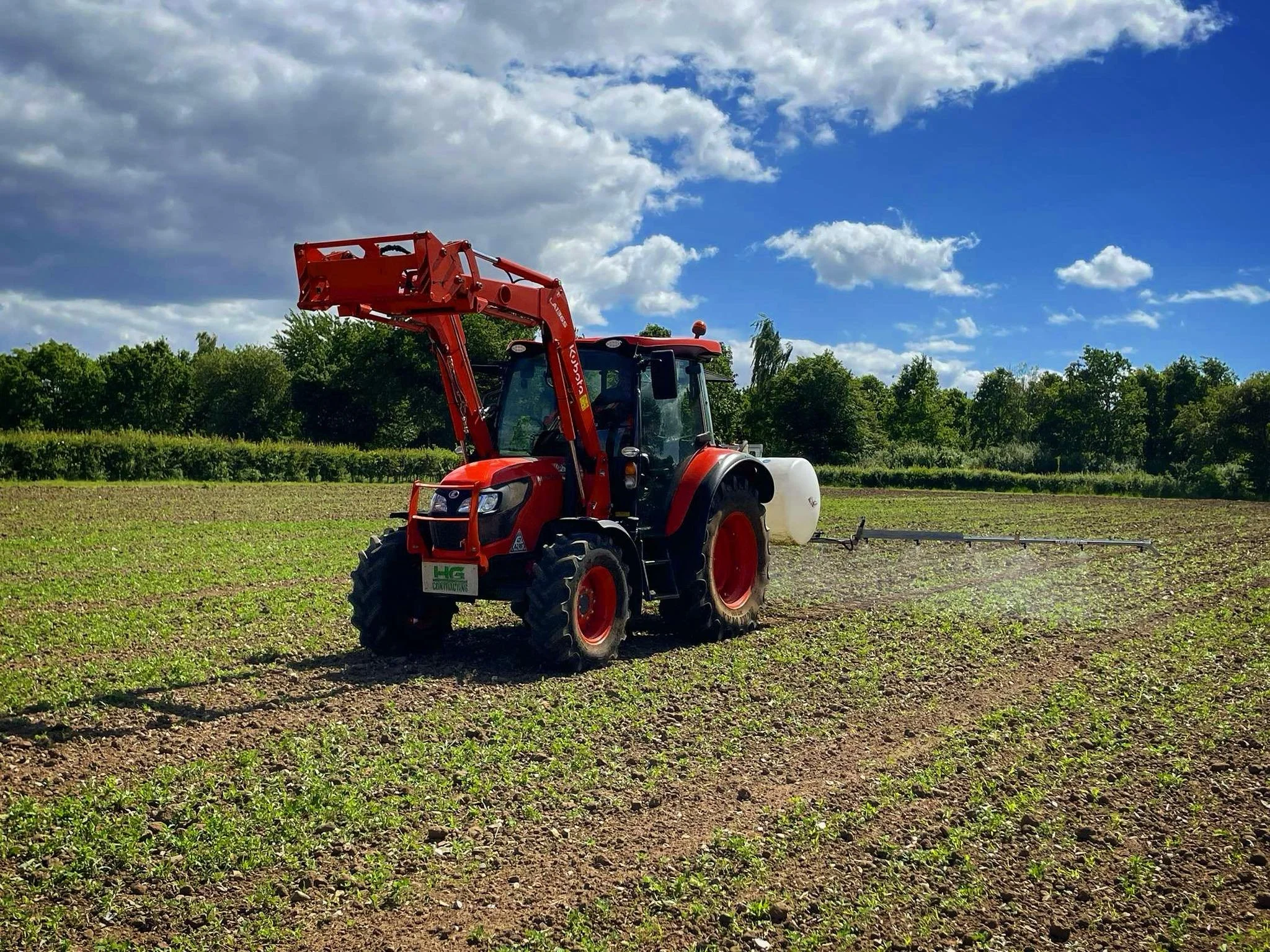 Red tractor spraying fertilizer or pesticide on a farm field during a sunny day with scattered clouds and green trees in the background.