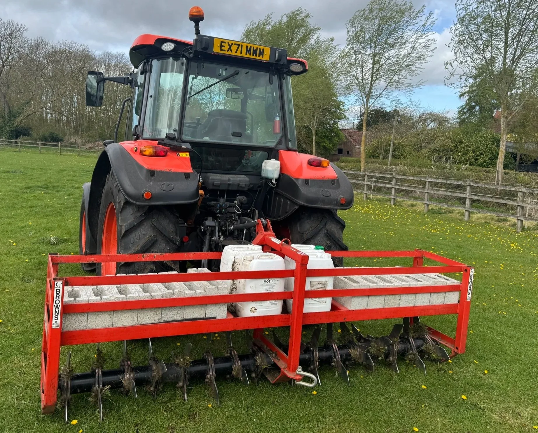 A red tractor with a mounted agricultural implement on a grassy field, surrounded by trees and a wooden fence, under a partly cloudy sky.