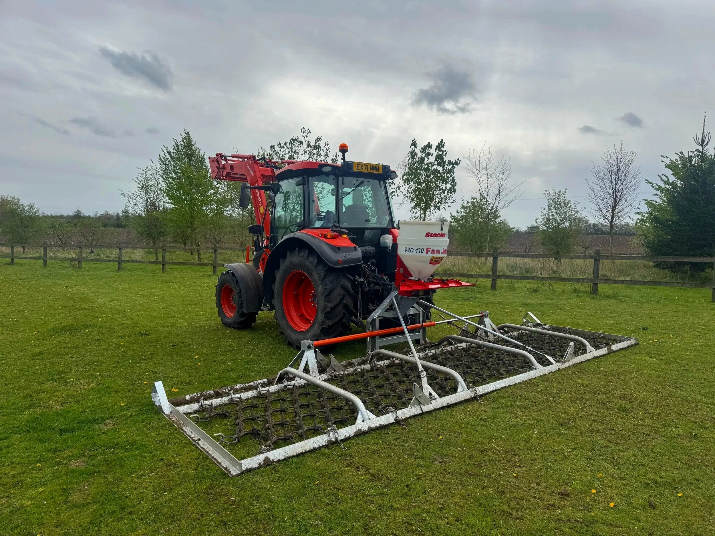 A red tractor with a large attachment working on a grassy field with trees and bushes in the background.