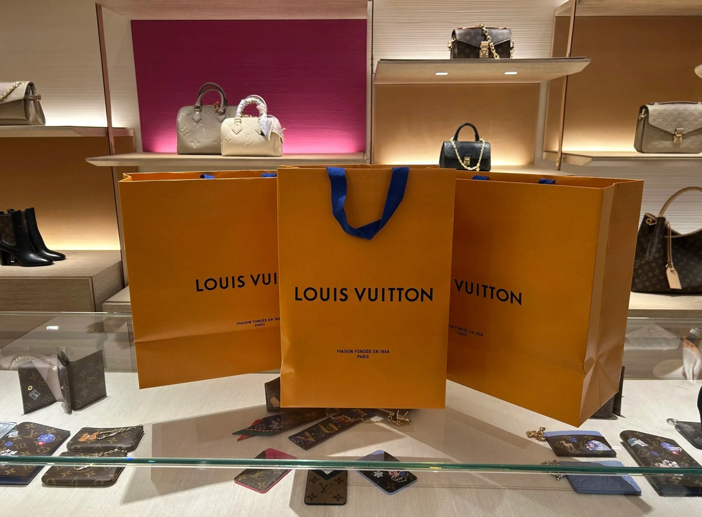 Three orange Louis Vuitton shopping bags with blue handles on a display counter at a luxury store, surrounded by luxury handbags, wallets, and accessories.