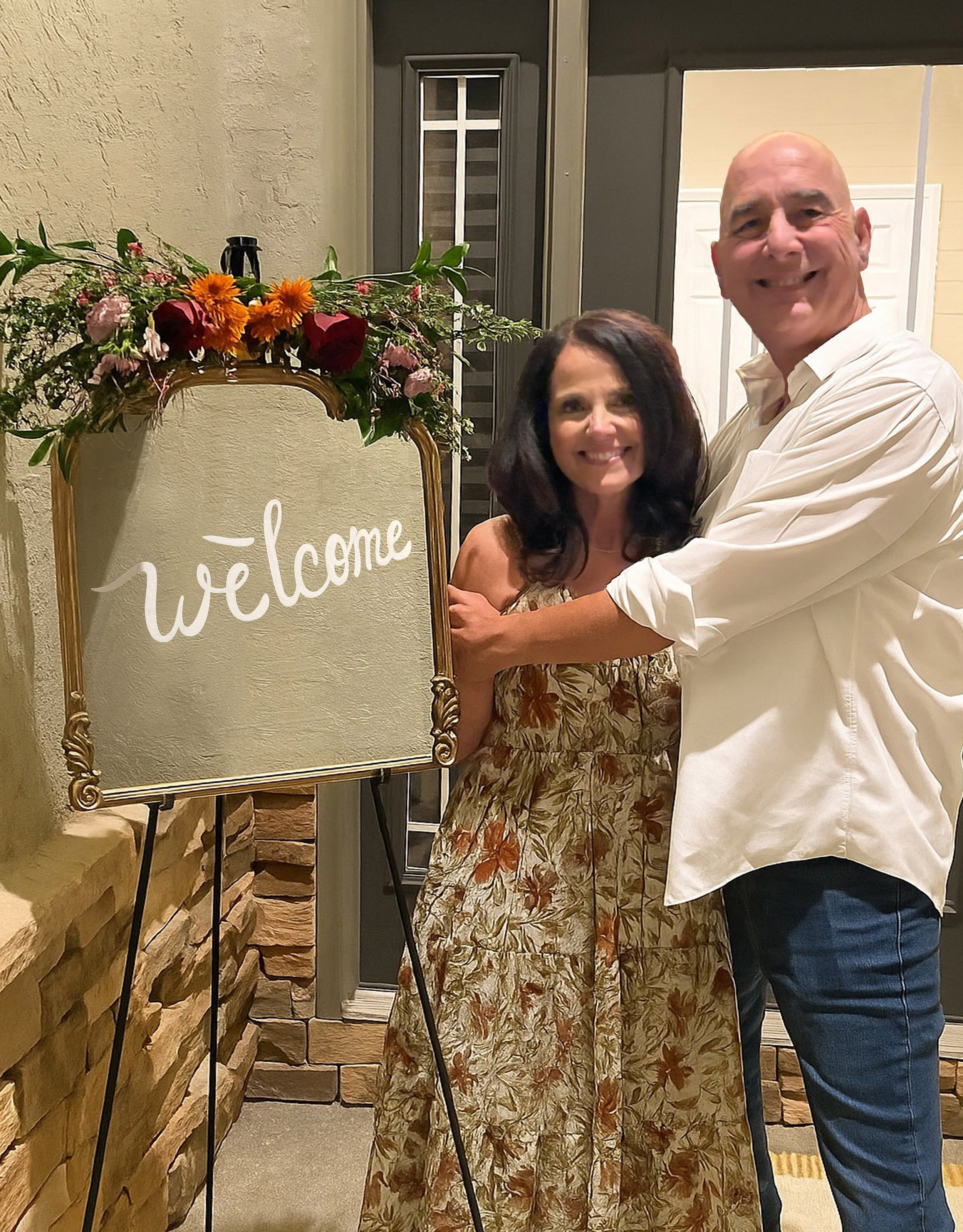 A smiling couple standing next to a decorative welcome sign with flowers on top, inside a building near a brick wall and door.