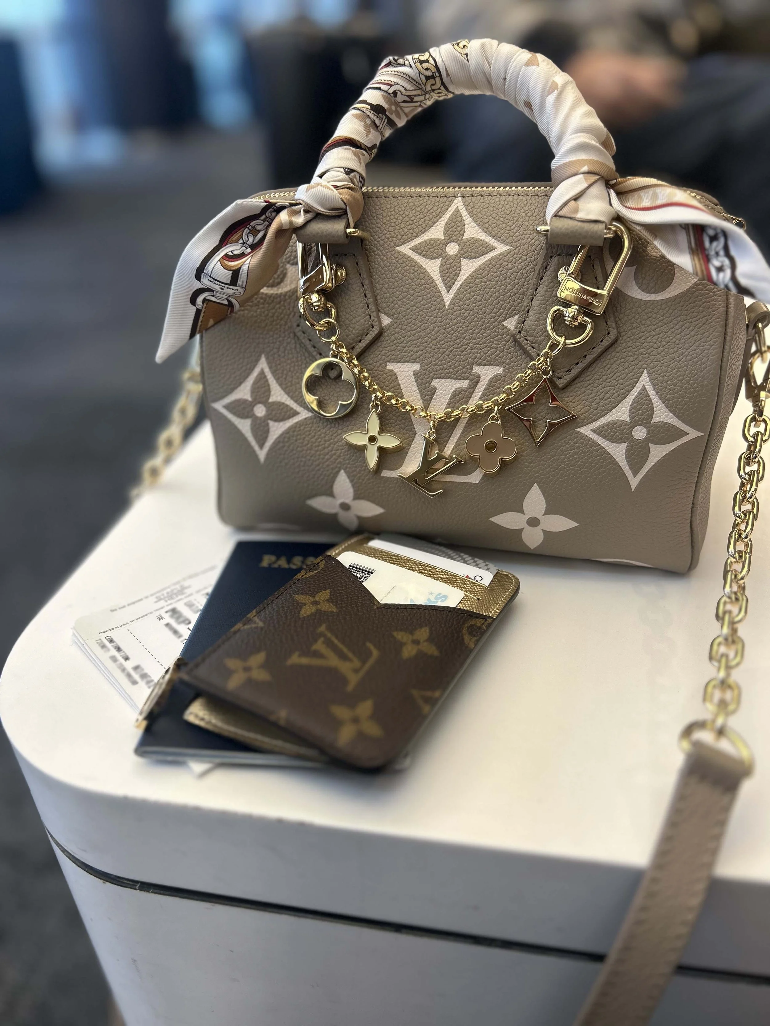 A beige Louis Vuitton handbag with white monogram print, decorated with a gold chain and charms, including a key and the LV logo. A matching scarf is wrapped around the handle. A Louis Vuitton wallet and a passport are placed on a white table.