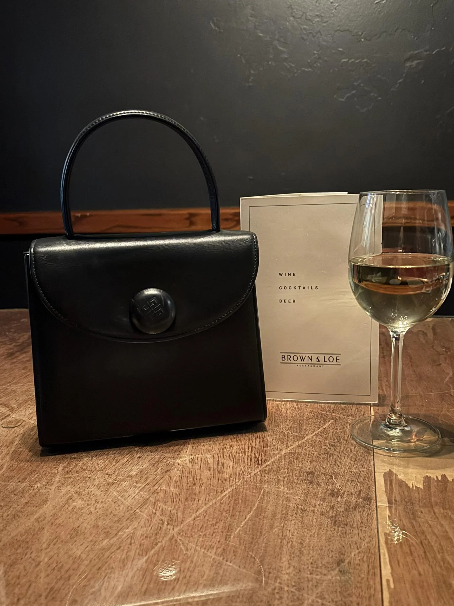 A black handbag, a menu for Brown & Loe restaurant, and a glass of white wine on a wooden table.